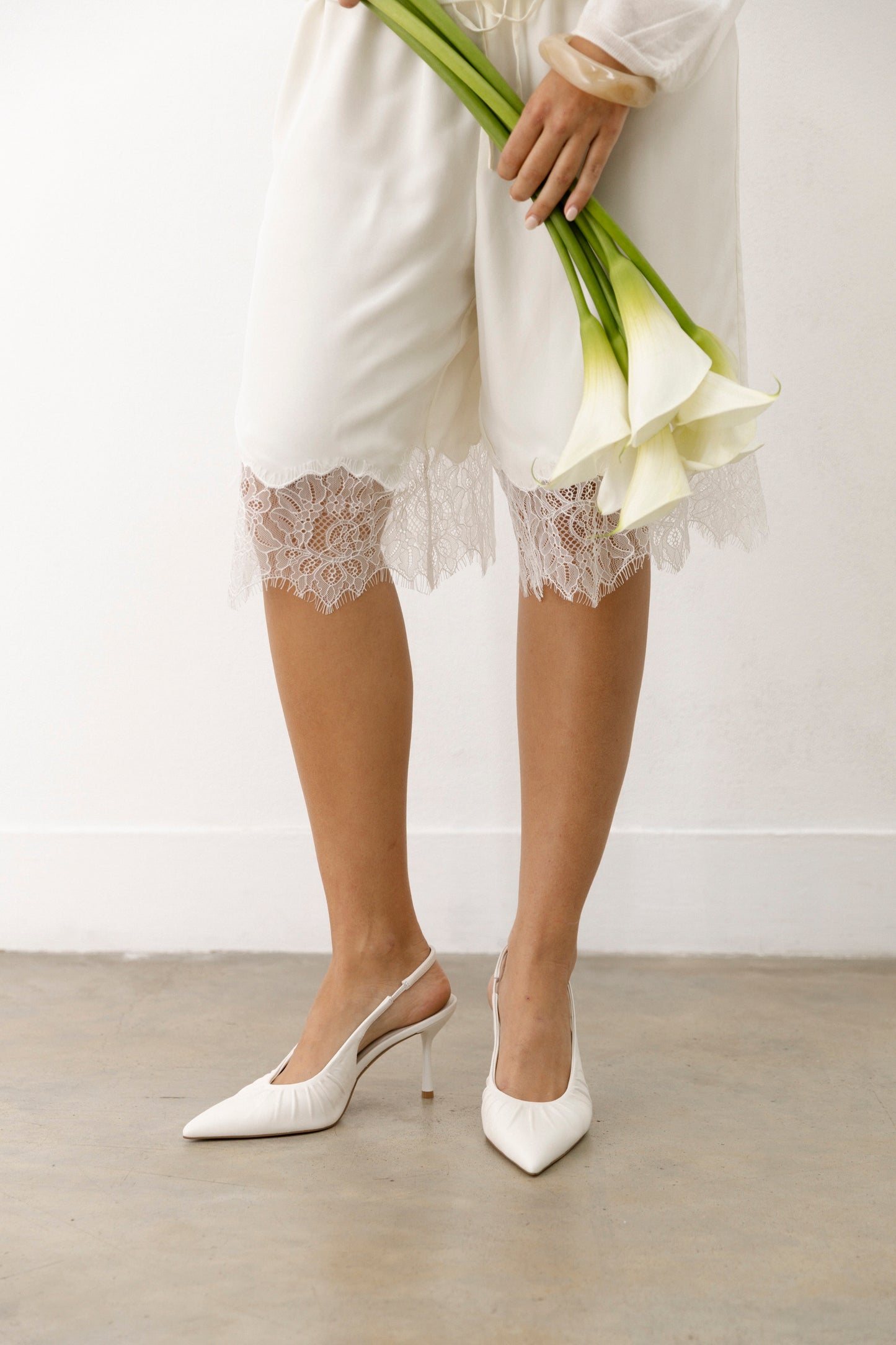 Wearing white leather slingbacks, a person in white lace-trimmed shorts stands on concrete, holding three white calla lilies.