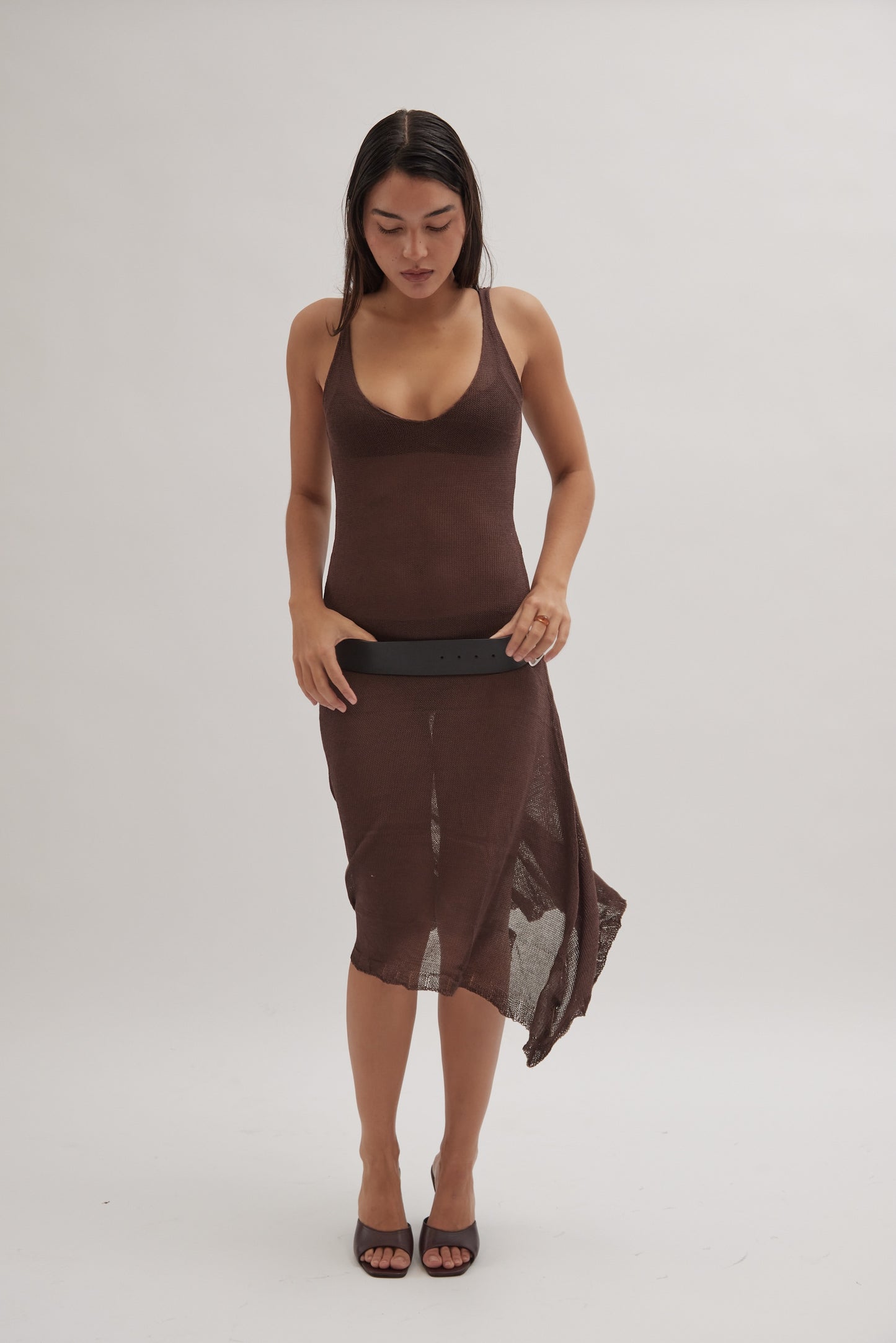 A woman in a sleeveless, brown, semi-sheer dress holds a black belt while wearing Realm's black leather mules, set against a plain light background.