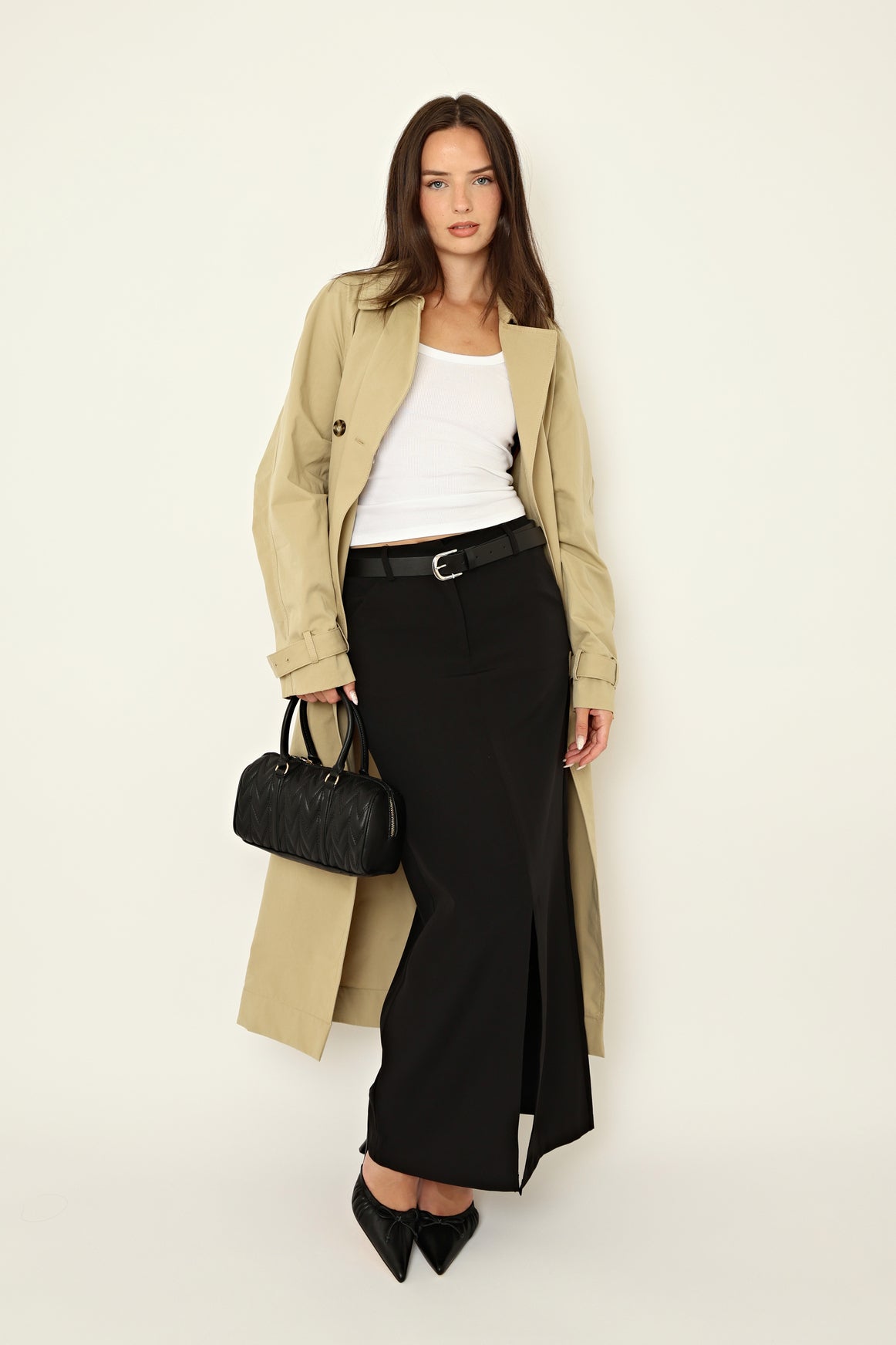 A woman stands against a plain light background, holding the Realm The Courts | in Black Leather small black handbag, styled with a beige trench coat, white tank top, black belted skirt with front slit, and event-ready heels.