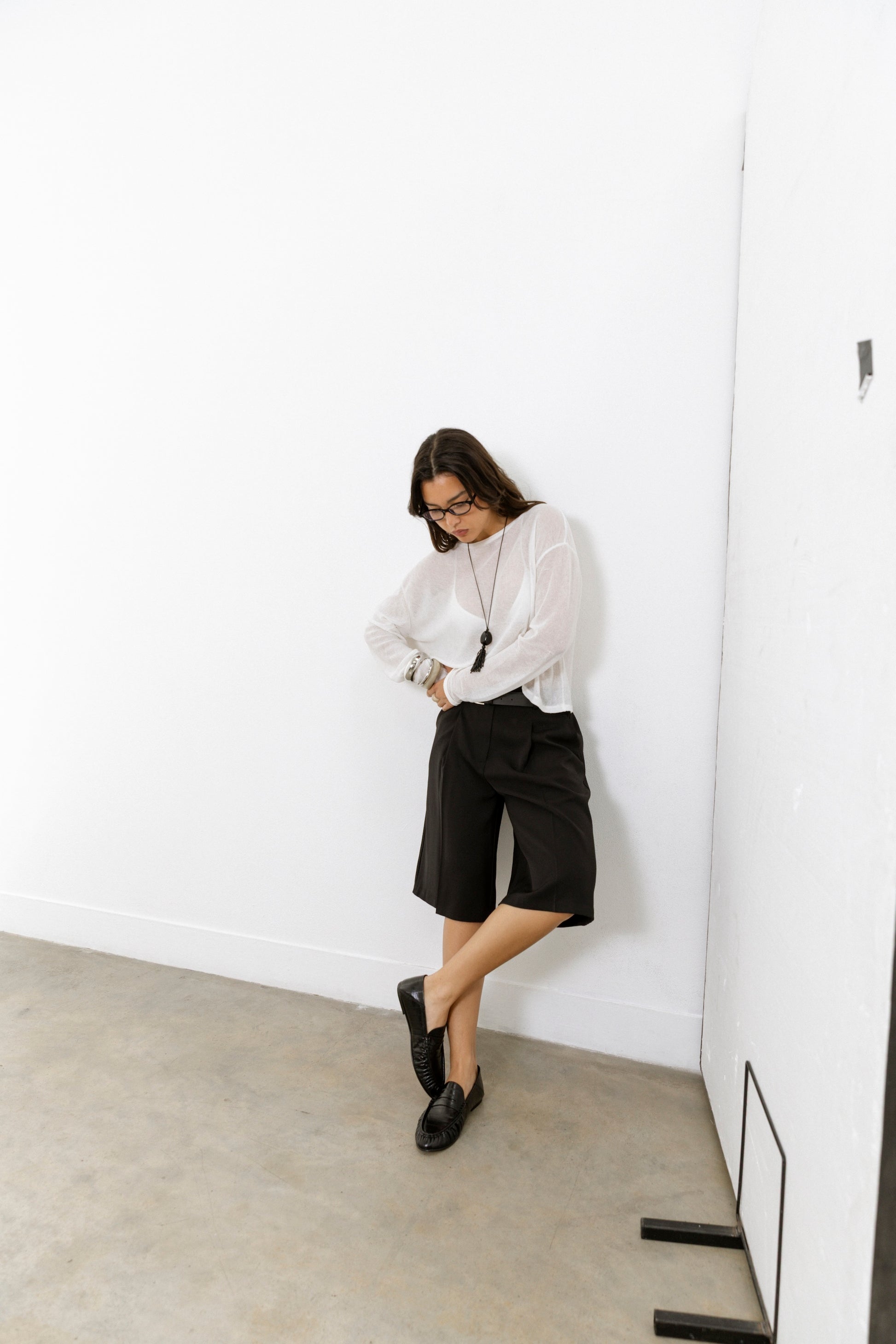 A person wearing a white long-sleeve shirt, black knee-length shorts, and black leather loafers stands in a minimal white-walled room, looking down with arms crossed and hands tucked into their sleeves.