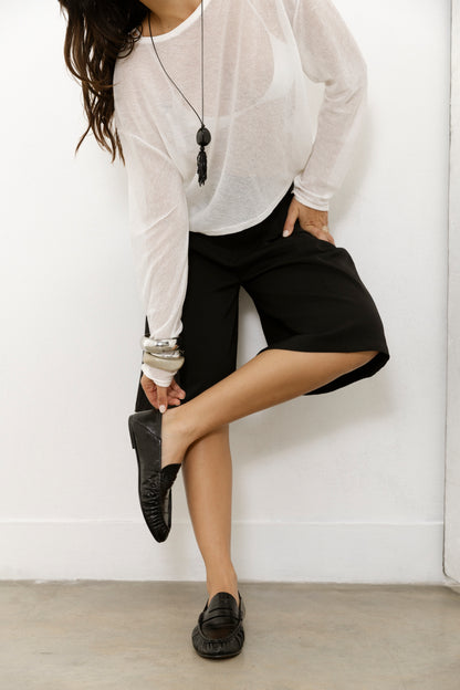 Wearing a sheer white long-sleeve top, black shorts, and jewelry, a person adjusts their Realm Form Loafers in black leather. They stand against a white wall on a concrete floor, with their face out of view.