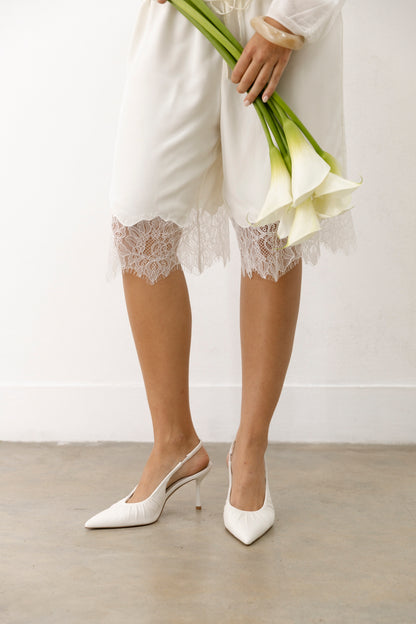 Wearing white leather slingbacks, a person in white lace-trimmed shorts stands on concrete, holding three white calla lilies.