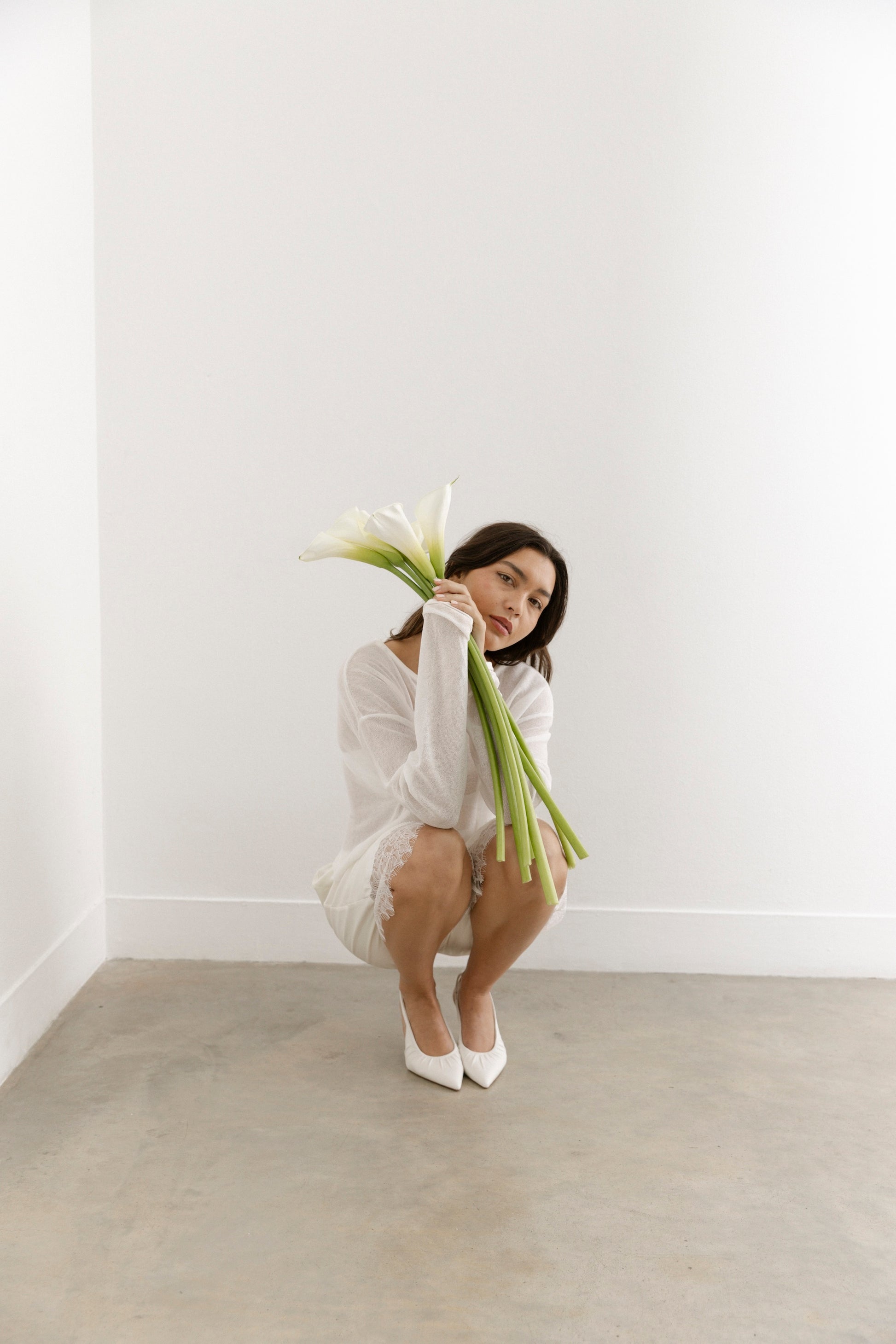 In a minimal, light-filled room, a woman in white crouches with white calla lilies, white  leather slingbacks peeking beneath her dress.