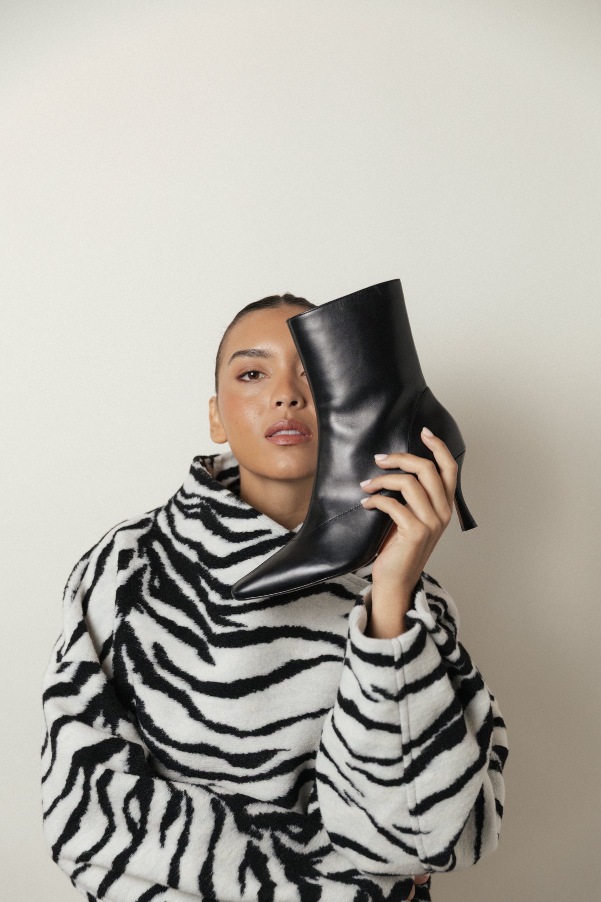 A person in a zebra-print coat holds the black leather Realm Encore Ankle Boot up to their face, standing against a plain light background—a perfect inspiration for adding these stylish black leather ankle boots to your capsule wardrobe.