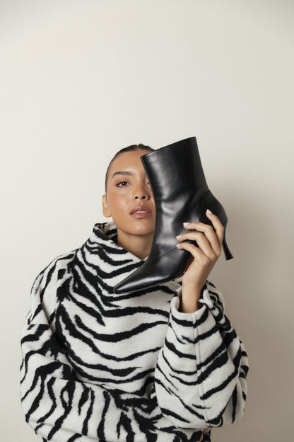 A person in a zebra-print coat holds the black leather Realm Encore Ankle Boot up to their face, standing against a plain light background—a perfect inspiration for adding these stylish black leather ankle boots to your capsule wardrobe.