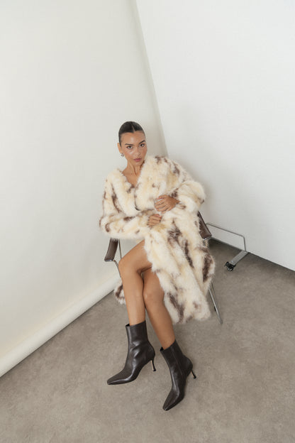 A woman sits on a chair with her legs crossed, exuding relaxed confidence in the black leather ankle boots. Her slicked-back hair and stylish cream and brown fur coat perfectly complement the minimal setting, capturing the effortless sophistication of a capsule wardrobe.
