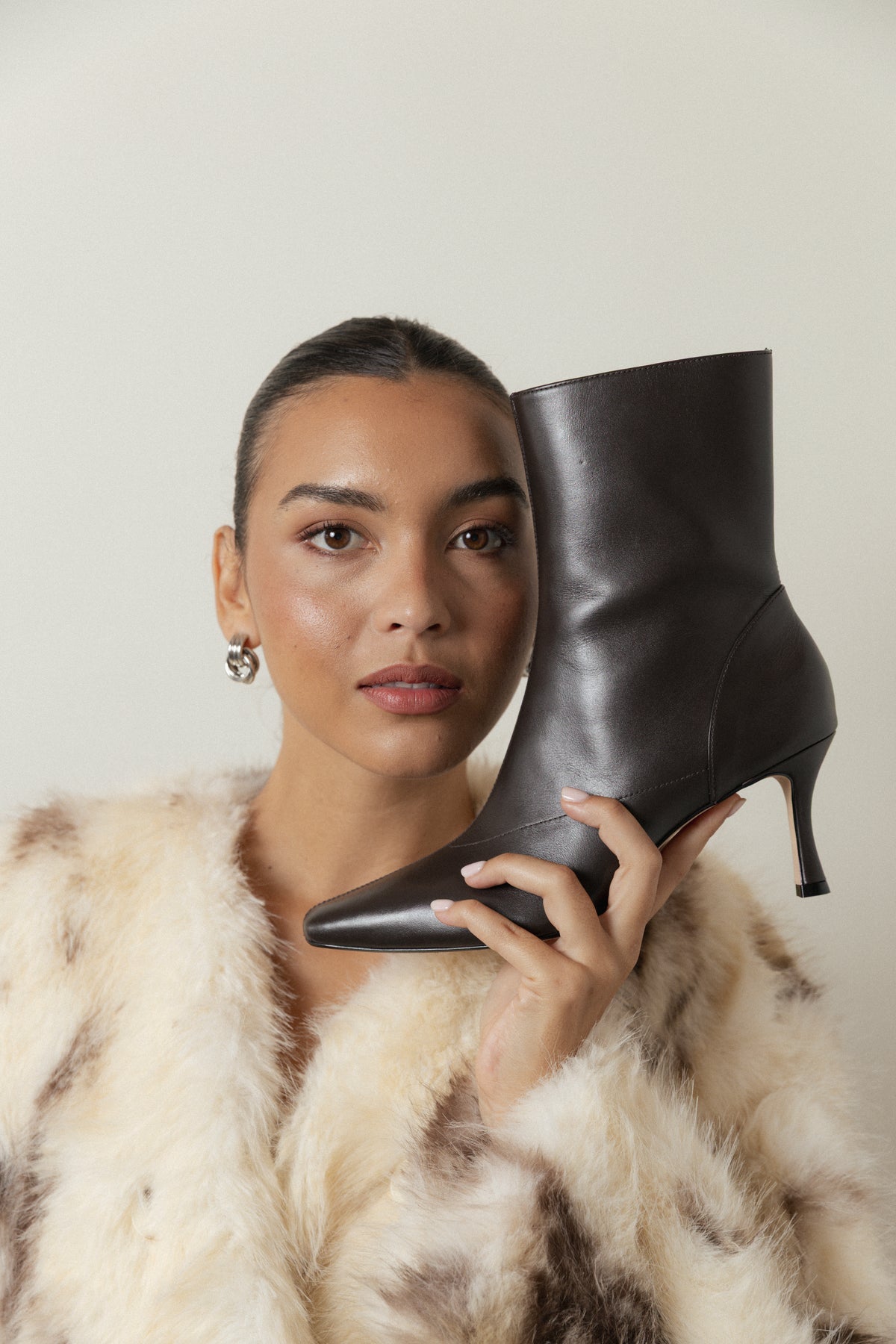A woman in a faux fur coat holds a pair of black leather ankle boots beside her face, gazing directly at the camera against a simple backdrop.