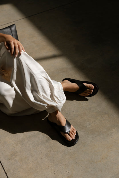 A person wearing white pants and black leather flats sits on a sunlit concrete floor, their left hand resting on their knee as sunlight casts distinct shadows across the surface.