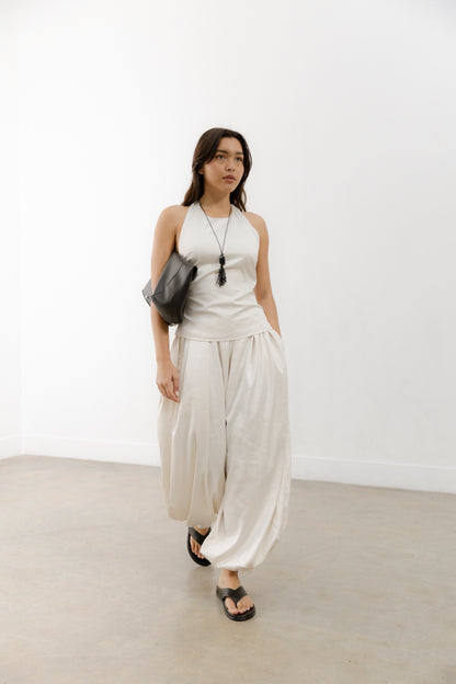 A woman in a sleeveless white top and loose pants walks in a minimalist room, wearing black leather flats, a long necklace, and a black shoulder bag.