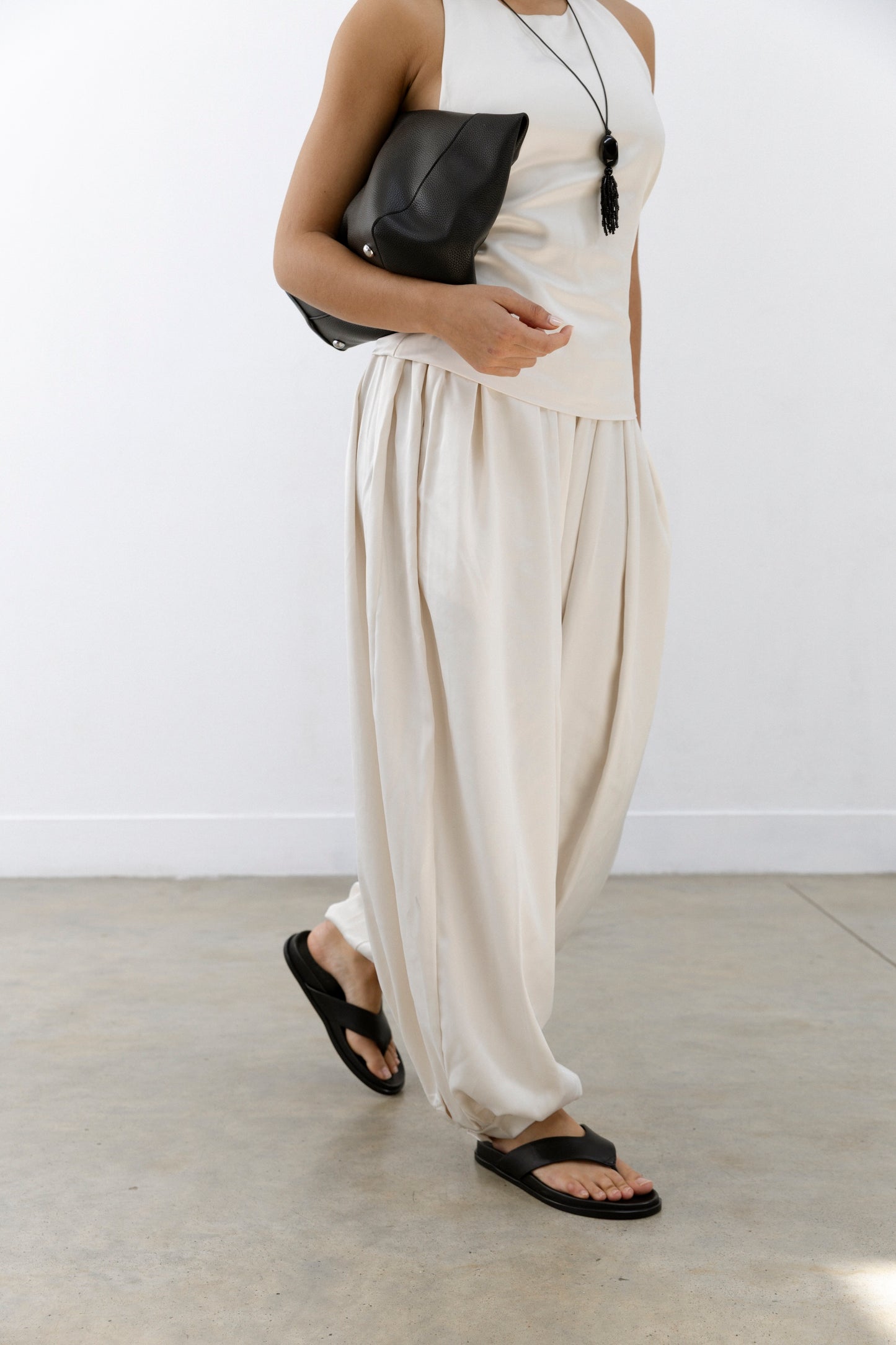 Wearing loose cream pants, a sleeveless beige top, and black leather flats, this person holds a black clutch on a light floor.