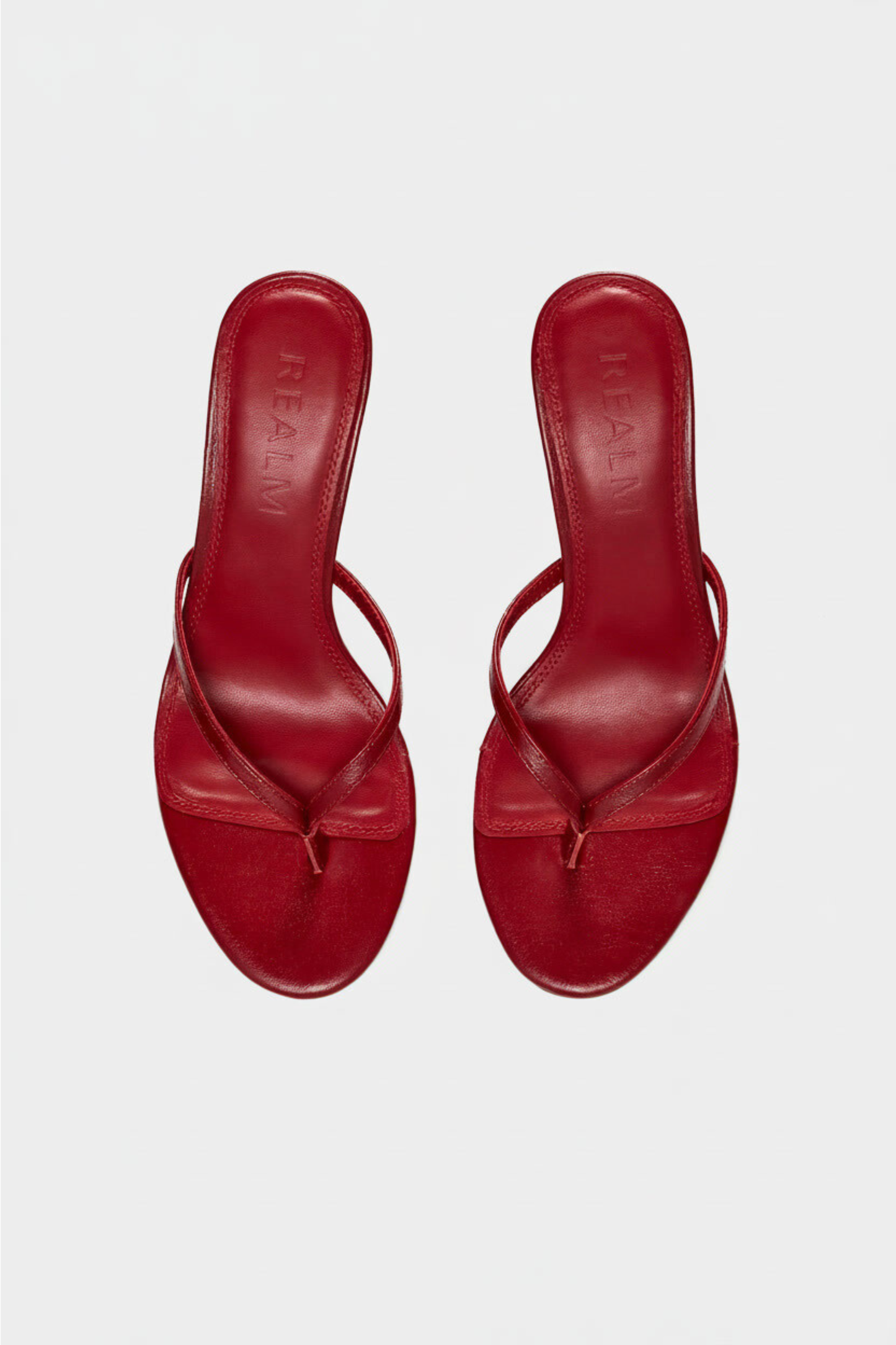 The Spirit Heeled Sandals by Realm in red leather feature thin straps, a toe thong design, and a slight heel on a white background.
