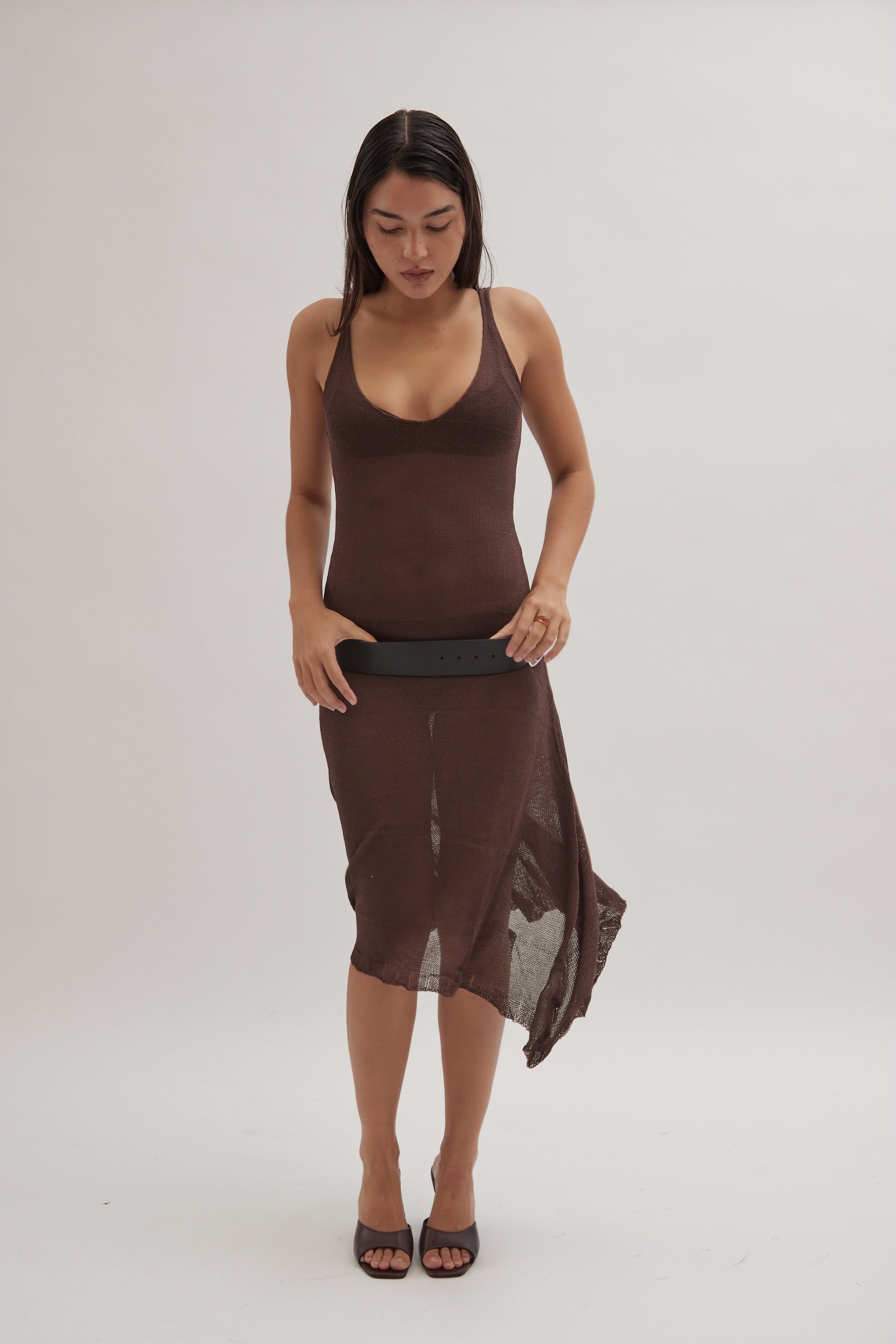 A woman in a sleeveless, brown, semi-sheer dress holds a black belt while wearing Realm's black leather mules, set against a plain light background.