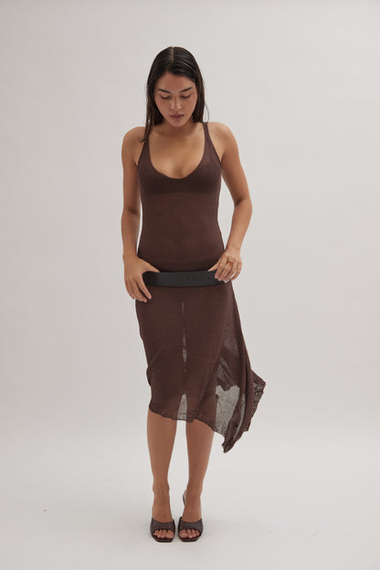 A woman in a sleeveless, brown, semi-sheer dress holds a black belt while wearing Realm's black leather mules, set against a plain light background.