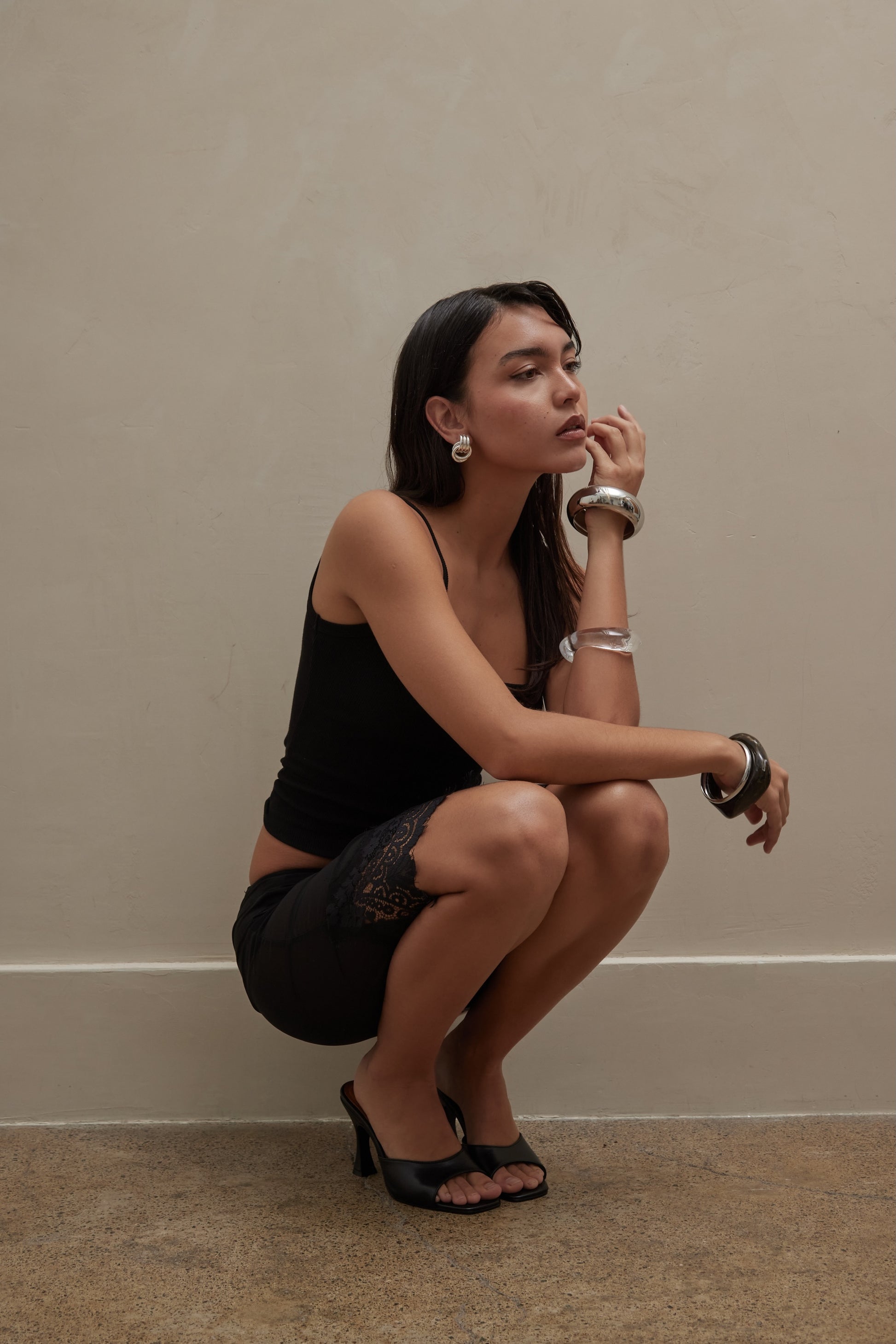 A woman in a black tank and lace-trim shorts, chunky silver jewelry, and black leather mules crouches by a beige wall, looking thoughtful.