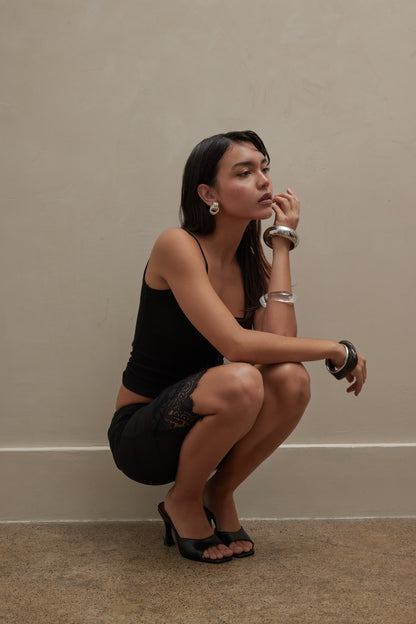 A woman in a black tank and lace-trim shorts, chunky silver jewelry, and black leather mules crouches by a beige wall, looking thoughtful.