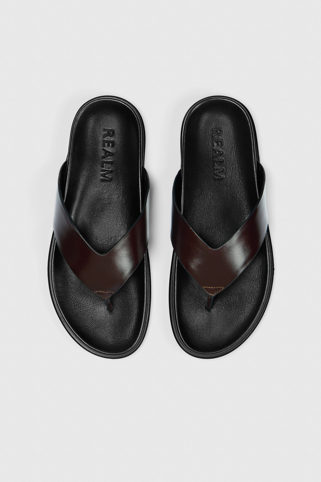 A pair of black leather Contour Flats by Realm, featuring crisscross straps, a contoured footbed, Realm embossed on the footbeds, displayed from above on a plain white background.