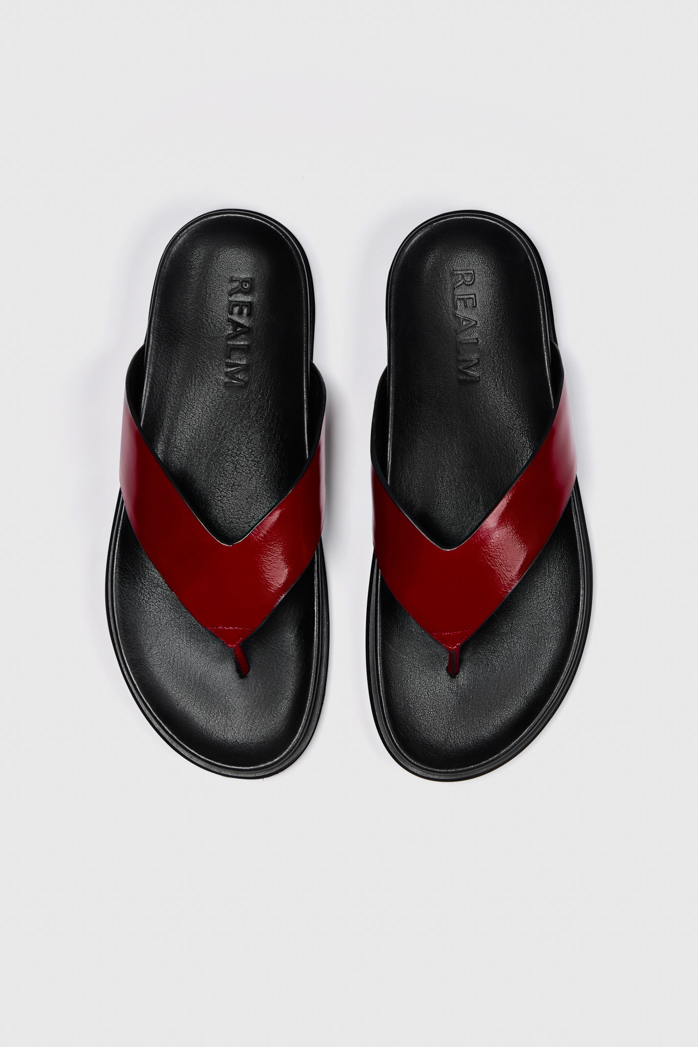 A pair of black leather Realm Contour Flats with wide, glossy red leather straps crossing over the top, on a plain white background. "Realm" is embossed on the contoured footbed of each sandal.