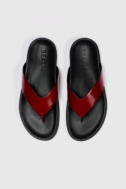 A pair of black leather Realm Contour Flats with wide, glossy red leather straps crossing over the top, on a plain white background. "Realm" is embossed on the contoured footbed of each sandal.