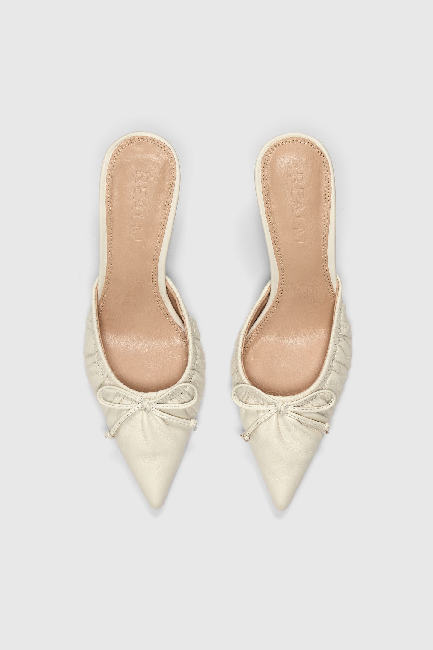 The Courts by Realm are vintage white leather shoes with pointed toes, ruched detailing, and small bows on the front, displayed from above on a light gray background.