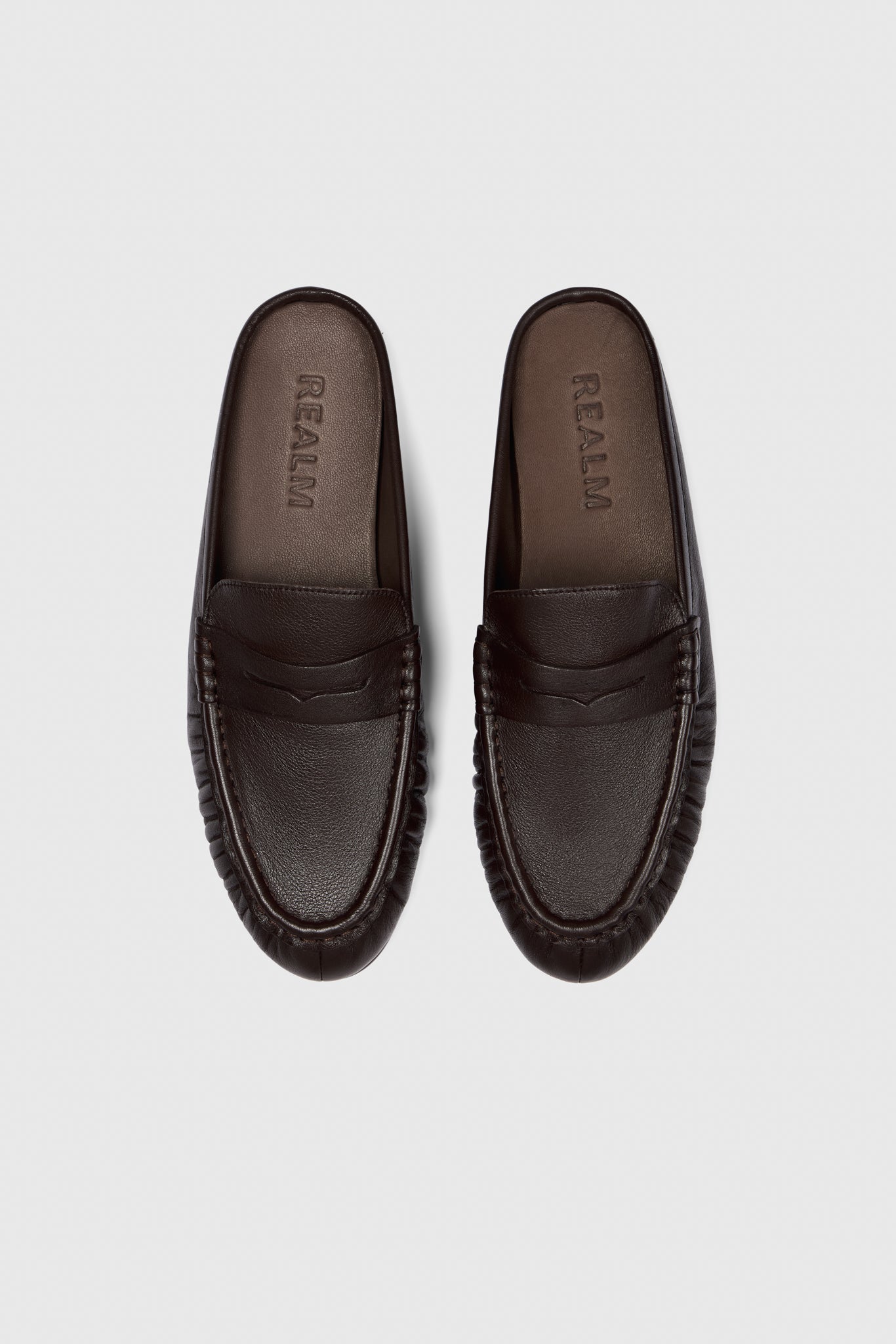 A pair of black leather Realm Everyday Loafers with rounded toes and stitched details are placed side by side on a light gray background. The insoles are embossed with the brand name.