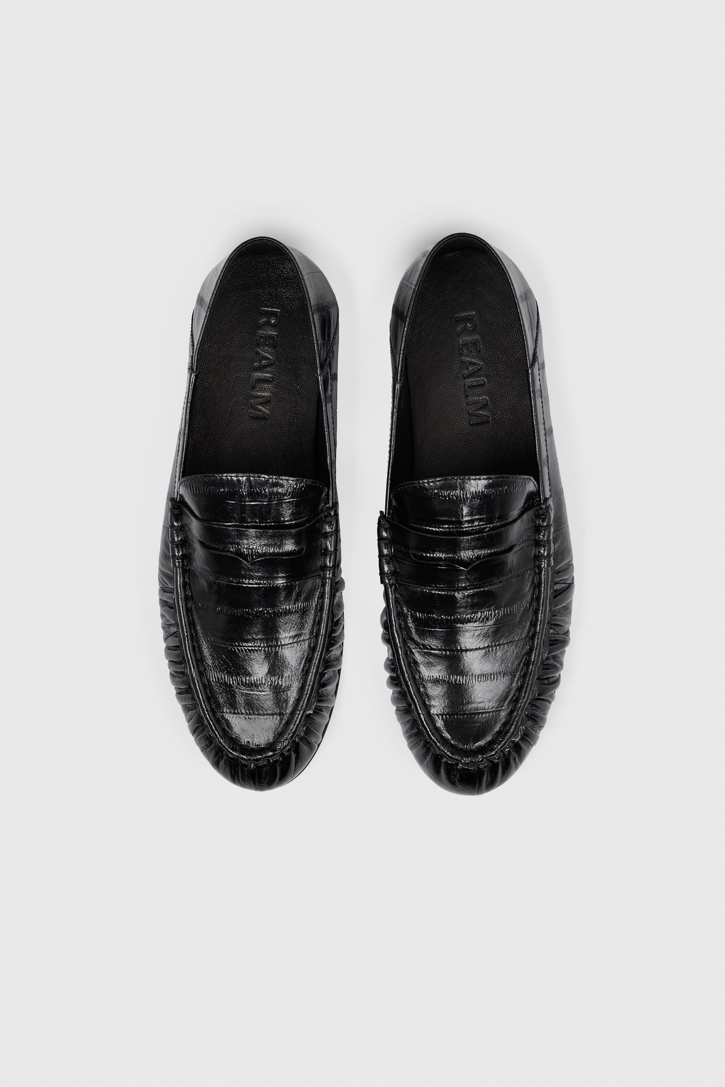 A pair of Realm’s Form Loafers in glossy black leather, shown from above on a light gray background. Embossed leather with toe stitching and the brand name visible inside.