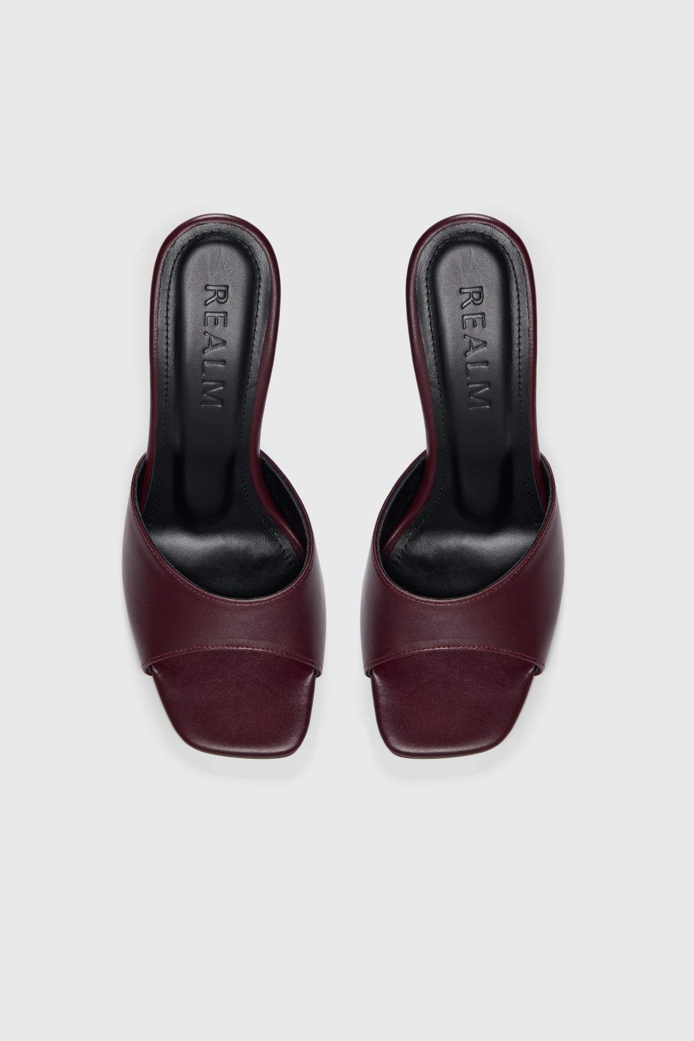 Slip-on burgundy leather mules with open toes, flared heels, and black insoles labeled "Realm," set against a light gray background.