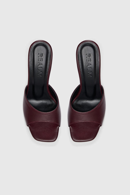 Slip-on burgundy leather mules with open toes, flared heels, and black insoles labeled "Realm," set against a light gray background.