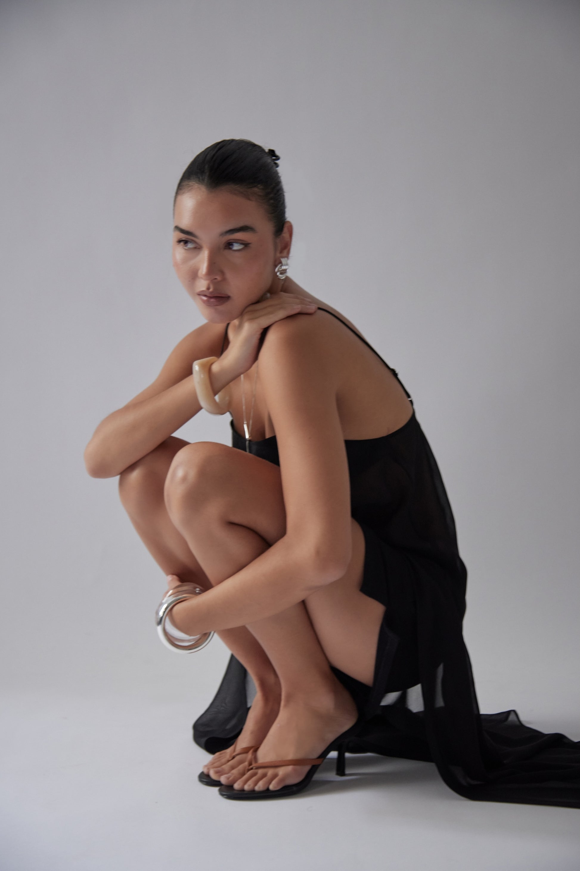 A woman with dark hair in a bun, in a sheer black dress and silver jewelry, squats wearing tan leather heeled sandals.