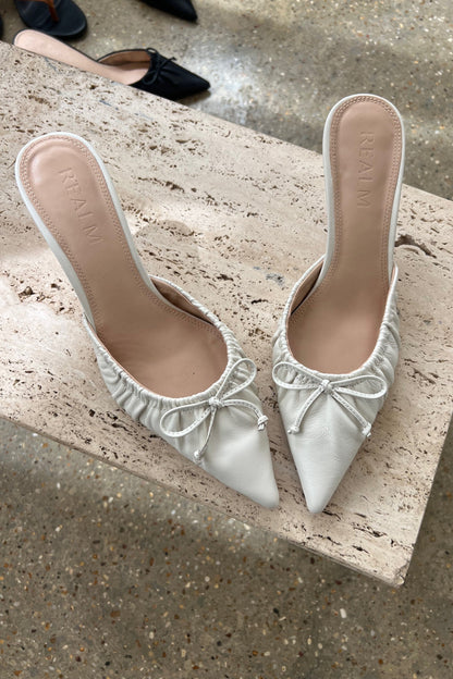 A pair of black leather heels by Realm with pointed toes, gathered detailing, and small front bows sits on a beige marble surface.