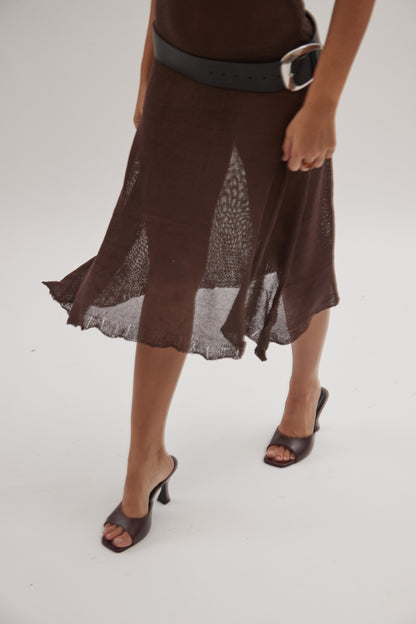 A person wearing a brown sheer skirt and black leather mules walks on a white surface. Only their lower body is visible, with one hand resting on the skirt.