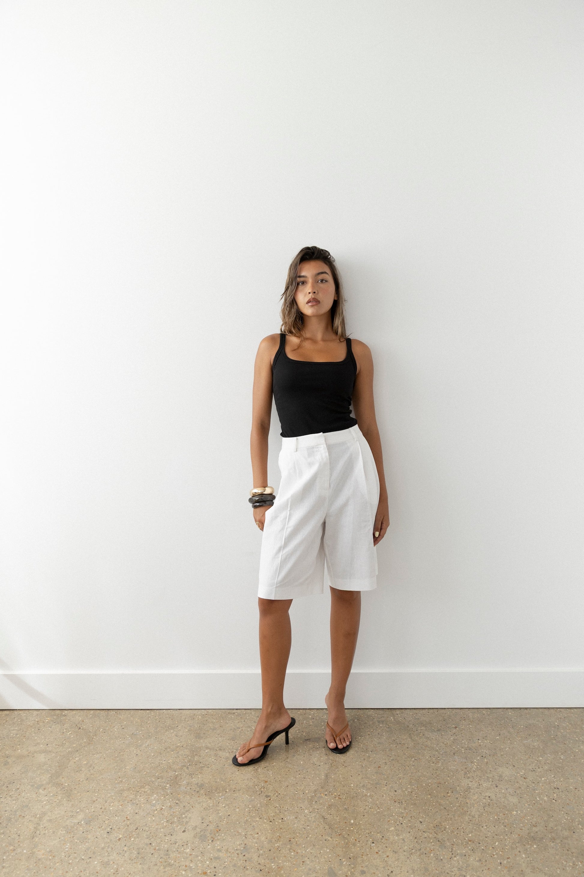 A woman stands against a white wall in a black sleeveless top, white knee-length shorts, and tan leather heeled sandals. Holding sunglasses in one hand, she gazes confidently at the camera.