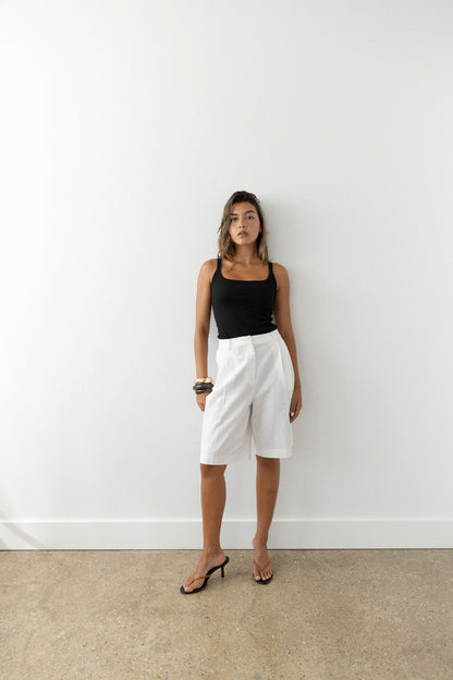 A woman stands against a white wall in a black sleeveless top, white knee-length shorts, and tan leather heeled sandals. Holding sunglasses in one hand, she gazes confidently at the camera.