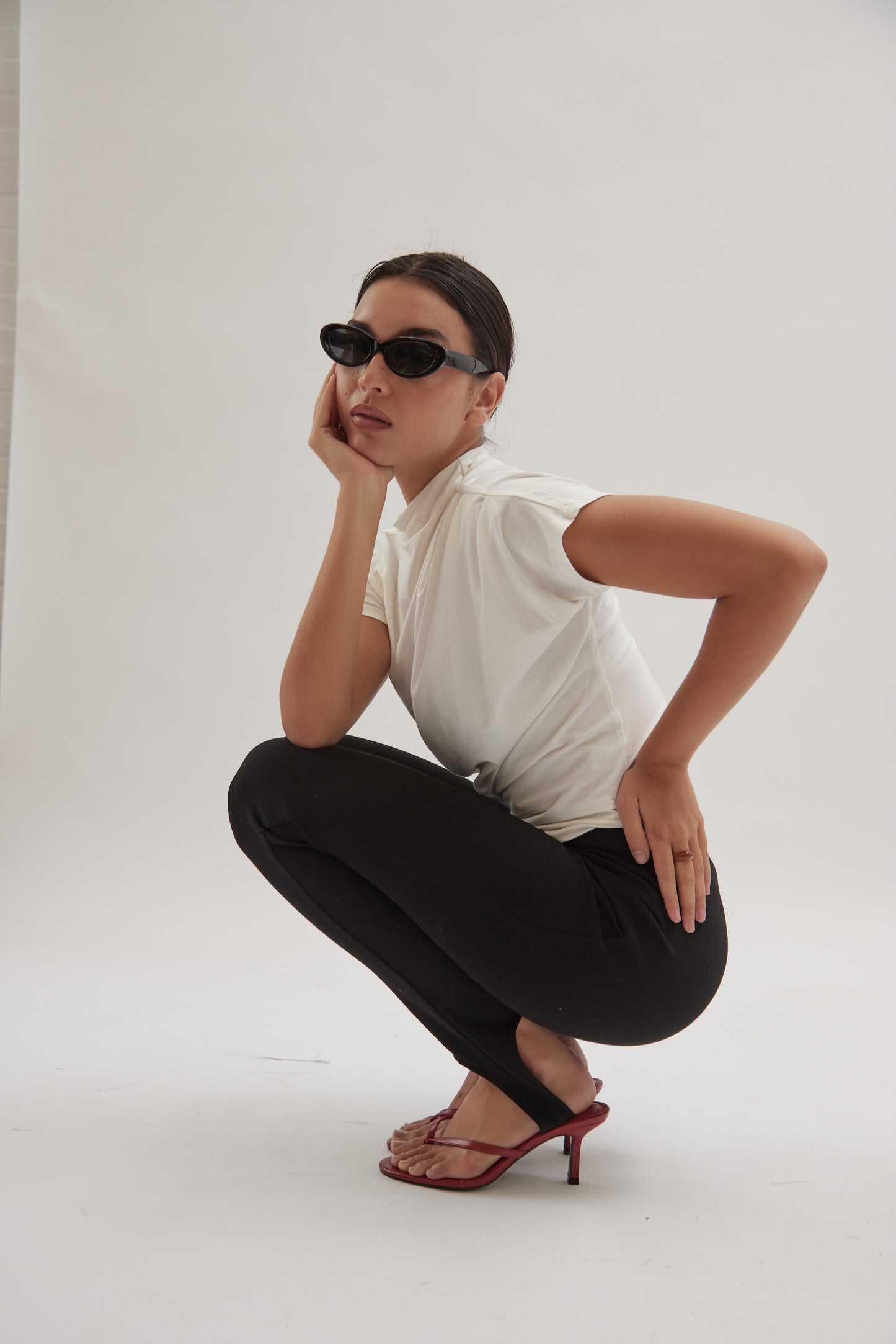 A person squats on the floor against a plain white background, wearing black leather heeled sandals, sunglasses, a white t-shirt, and black pants.