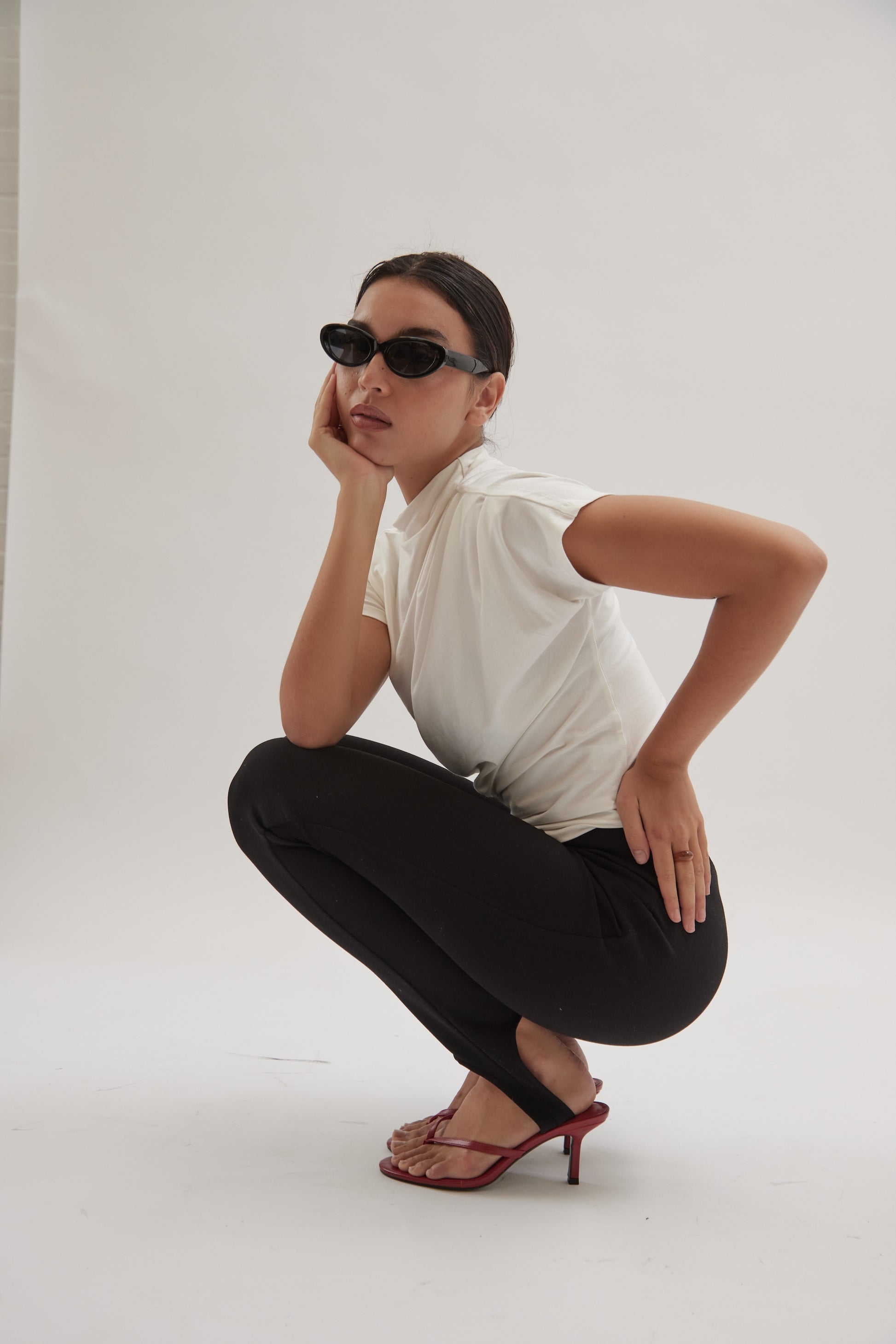 A person squats on the floor against a plain white background, wearing black leather heeled sandals, sunglasses, a white t-shirt, and black pants.